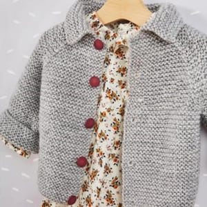 May include: A gray knit baby cardigan with a collared neckline and red buttons. The cardigan is layered over a floral print dress. The outfit is hanging on a wooden hanger.