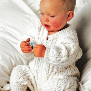 以下が含まれることがあります： ケーブル編みのパターンが付いた白いニットのベビーロンパース。ロンパースは、白い毛布の上に座っている赤ちゃんが着用しています。