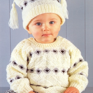 Puede incluir: Un suéter de punto blanco con un patrón de rombos negro y gris. El suéter tiene mangas largas y cuello redondo. El suéter lo lleva puesto un niño que lleva un gorro de punto blanco con detalles negros y grises y borlas.