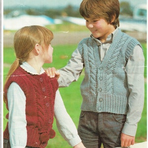 Może przedstawiać: Dwoje dzieci w dzianinowych kamizelkach. Dziewczynka ma na sobie czerwoną kamizelkę z wzorem warkocza, a chłopiec niebieską kamizelkę z wzorem warkocza. Obie kamizelki mają guziki z przodu.