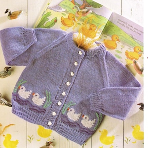 May include: A purple knitted baby cardigan with a white button fastening and embroidered ducks on the front. The cardigan is laid out on a white surface with a children's book in the background.