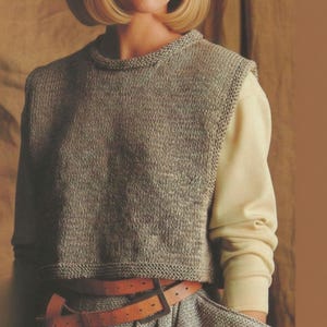 May include: A knitted, sleeveless sweater in a neutral gray color. The sweater has a crew neck and a ribbed hem. A light-colored long-sleeved shirt is worn underneath. A brown leather belt is visible at the waist.