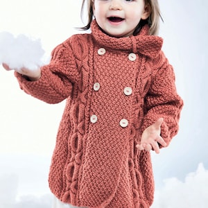 May include: A coral-colored, double-breasted knitted coat with large wooden buttons. The coat features a high collar and cable knit detailing. The child is holding a white fluffy object. The coat is a warm and cozy garment.
