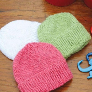 May include: Three knitted baby hats in white, pink, and green. The hats are made of a soft yarn and have a ribbed brim. The hats are laying on a wooden surface.