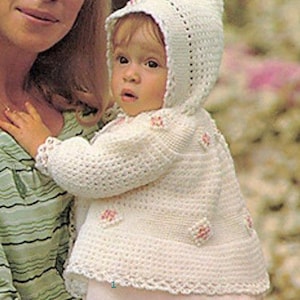 Könnte beinhalten: Ein weißes gehäkeltes Baby-Jäckchen mit Kapuze und rosa Blumenapplikationen. Das Jäckchen hat einen zarten Häkelrand an den Kanten.