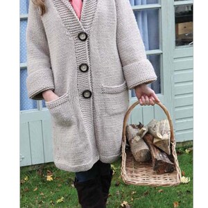 May include: A woman wearing a long, light beige knitted cardigan with a pink scarf and brown boots. She is holding a wicker basket filled with logs.