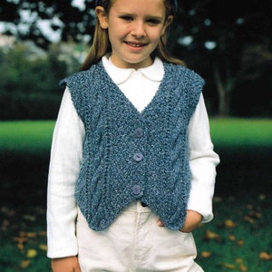 May include: A young person wearing a white long-sleeved shirt with a blue knitted vest and white pants. The vest has a V-neck and buttons down the front. The person is wearing a blue baseball cap with the word "Maypole" on it.