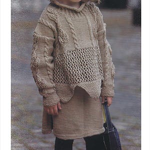 May include: A young girl wearing a brown knitted sweater and skirt with a matching brown knitted hat. She is holding a dark blue handbag.