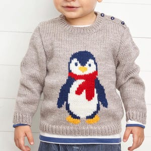 Op de afbeelding: Een lichtbruine gebreide trui met een pinguïnontwerp. De pinguïn heeft een blauw lichaam, een witte buik, een rode sjaal en gele voeten. De trui heeft blauwe knopen op de schouder en blauwe boorden aan de manchetten en zoom.