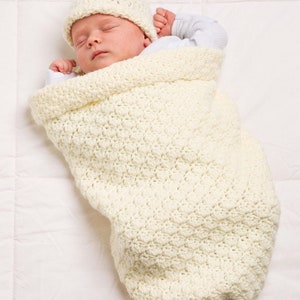 May include: A white crocheted baby swaddle blanket with a white crocheted hat. The baby is sleeping peacefully inside the swaddle.
