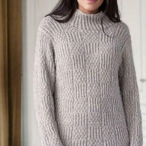 May include: A light gray, long-sleeved sweater with a high, ribbed collar. The sweater features a textured, geometric pattern across the body and sleeves. The model is wearing the sweater.