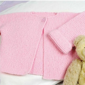 May include: A pink knitted cardigan with a button fastening. The cardigan is laid out on a white surface.