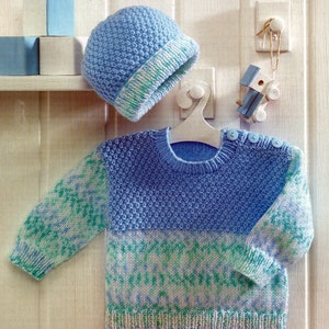 May include: A blue knitted baby sweater with a green and white patterned design. The sweater has a round neckline and long sleeves. A matching blue knitted hat is also included.