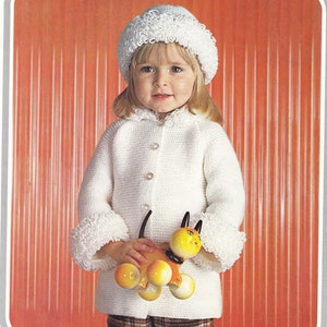 May include: A white knitted cardigan with a white crocheted collar and cuffs. The cardigan has a button closure and is worn by a child holding a yellow toy dog.