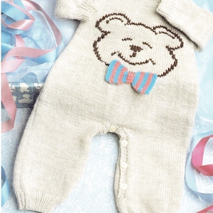 May include: A cream-coloured knitted baby romper with a brown bear face and a blue and pink striped bow tie. The romper has long sleeves and legs, with button fastenings down the front.