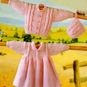 May include: A pink knitted baby dress and cardigan set with a matching hat. The set features a cable knit pattern and button closures.