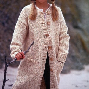 Puede incluir: Cárdigan largo de punto beige con grandes botones de madera y dos bolsillos delanteros. El cárdigan lo lleva una persona con coletas rubias, sosteniendo un pequeño palo. El cárdigan es de color neutro y parece hecho a mano.