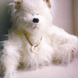 Può includere: Un orsacchiotto bianco e soffice fatto a mano, con un naso nero e piccoli occhi scuri. L'orso ha una collana marrone chiaro con un ciondolo di legno. L'orso è sdraiato, con le zampe distese.