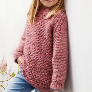 May include: A dusty rose-colored knitted sweater with a high neckline. The sweater has long sleeves and a textured knit pattern. The person is wearing blue jeans. The sweater appears to be handmade.