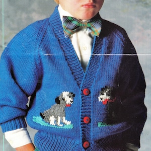 May include: A bright blue knitted cardigan with red buttons and dog appliqués. The cardigan features a V-neck and long sleeves. The dog designs are in shades of gray, white, and black, with red accents. The cardigan is worn over a white shirt and a plaid bow tie.