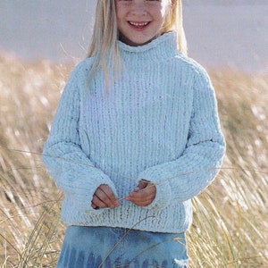 May include: A young girl wearing a light blue knitted sweater with a high neck and a denim skirt with embroidered details. She is standing in a field of tall grass.