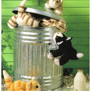 Könnte beinhalten: Drei gestrickte Katzenspielzeuge in einem silbernen Mülleimer mit einem teilweise geöffneten Deckel. Die Katzenspielzeuge sind braun-weiß, schwarz-weiß und orange-weiß. Neben dem Mülleimer liegen zwei Glasmilchflaschen auf dem Boden.