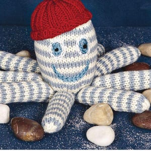 Könnte beinhalten: Ein handgefertigtes, gestricktes Oktopus-Spielzeug mit blau-weiß gestreiftem Körper, blauen Augen und einem lächelnden Mund. Es trägt eine rote Strickmütze. Das Spielzeug ist von kleinen, glatten Steinen auf einer blauen Oberfläche umgeben.