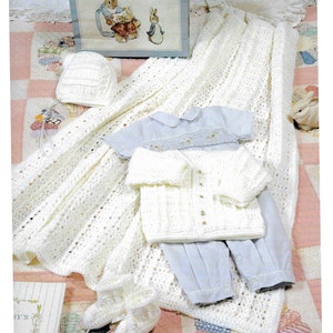 Może przedstawiać: Biały zestaw do chrztu dla niemowlęcia wykonany na szydełku, w skład którego wchodzi kocyk, czapka, buciki i kardigan. Zestaw ułożony jest na różowo-białej kołdrze z książką i ramką na zdjęciu w tle.