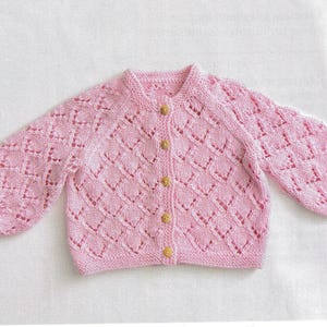 May include: A light pink, knitted baby cardigan with a diamond pattern. The long sleeves are gathered at the cuffs. The cardigan has a round neckline and is fastened with five wooden buttons.