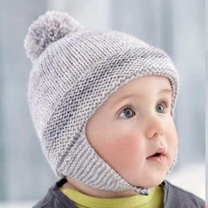 May include: A gray knitted baby hat with a pom pom on top and ear flaps. The hat has a darker gray stripe around the bottom.