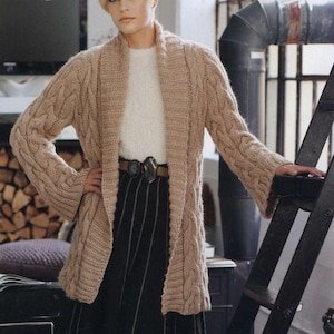 Op de afbeelding: Een lange, beige gebreide cardigan met een kabelbreipatroon. De cardigan heeft een brede, open kraag en lange mouwen. De cardigan wordt gedragen over een wit shirt en een zwarte rok.