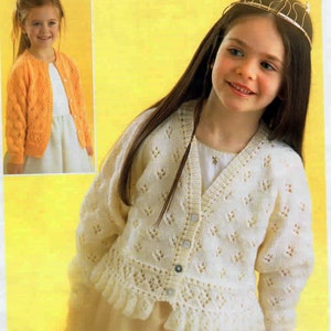 May include: A white knitted cardigan with a delicate lace pattern and a frilled hem. The cardigan has a V-neck and button fastening. The model is wearing a white dress and a crown.