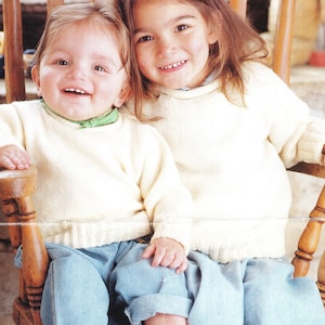May include: Two children wearing white knitted sweaters sit in a wooden rocking chair. The child on the left is wearing blue jeans and has a green scarf around their neck. The child on the right has long brown hair.