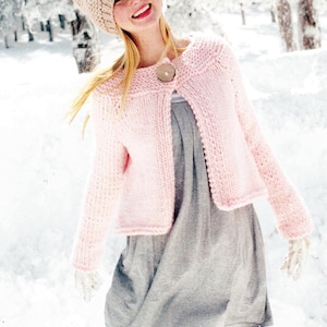 May include: A pink knitted cardigan with a single button closure and long sleeves. The cardigan is worn over a gray dress and the model is wearing a brown knit hat.