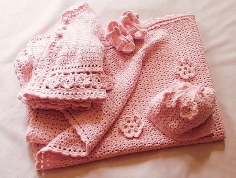 May include: A pink crocheted baby set including a blanket, a cardigan, a hat, and a pair of booties.