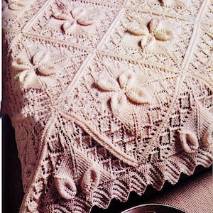 May include: A cream-colored, hand-knitted blanket with a floral pattern. The blanket features raised, three-dimensional flower motifs and a decorative, scalloped edge. The intricate design showcases detailed openwork and textured stitches, creating a cozy and elegant textile.