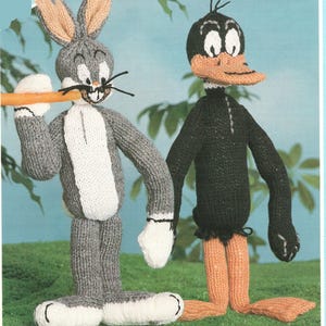 May include: Hand-knitted Bugs Bunny and Daffy Duck figures. Bugs Bunny is gray and white, holding a carrot. Daffy Duck is black with an orange beak and feet. Both figures are standing, with a light blue and green background.
