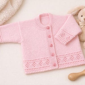 Peut inclure: Un gilet en tricot rose clair à manches longues avec une encolure ronde, cinq boutons assortis sur le devant et un motif ajouré en losanges texturé le long des poignets et de l'ourlet inférieur du vêtement.