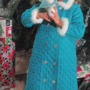 May include: A young girl wearing a blue knitted robe with white fur trim holds a white stuffed bunny. The robe has a button closure and is decorated with silver buttons. The girl is standing in front of a Christmas tree and wrapped presents.