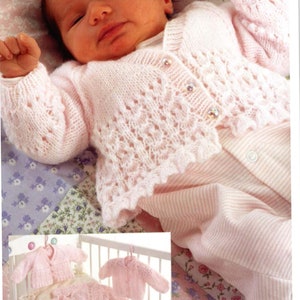 Op de afbeelding: Een roze gebreid babyvestje met een delicaat kantpatroon en parelmoerknopen. Het vestje is te zien op een baby die in een wiegje ligt.