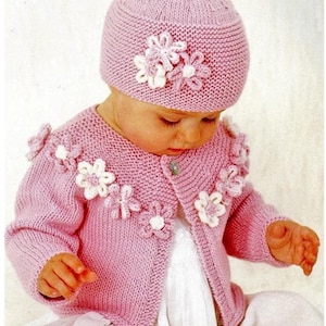 Pode incluir: Um casaco de tricô rosa para bebê com detalhes de flores brancas. O casaco tem fecho de botão e mangas compridas. O bebê também está usando um chapéu de tricô rosa com detalhes de flores brancas.