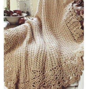 May include: A beige crocheted blanket with a scalloped edge. The blanket is draped over a chair, showcasing its intricate stitch pattern and openwork design. The color is a warm, neutral tone, suitable for various home decor styles.