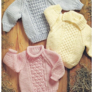 May include: Three knitted baby sweaters in different colours: light blue, cream, and pink. Each sweater has a different stitch pattern.