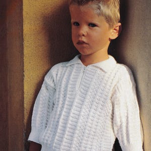 Op de afbeelding: Een kind draagt een witte gebreide trui met een kraag en een gedeeltelijke ritssluiting. De trui heeft een textuurpatroon. Het kind staat voor een muur. Het kind draagt een rood-witte geruite korte broek.