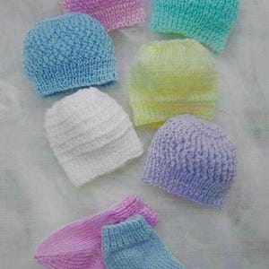 May include: A selection of hand-knitted baby hats and socks in pastel shades. The hats are in pink, blue, green, yellow, white, and purple. The socks are pink and blue. The items are displayed on a white surface.