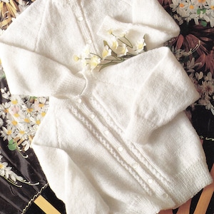 May include: Two white knitted cardigans with button fastenings. The cardigan on top has a round neckline and long sleeves. The cardigan on the bottom has a V-neckline and long sleeves.