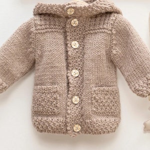 Op de afbeelding: Een bruin gebreid babyjasje met een capuchon, twee voorzakken en houten knopen. Het jasje is gemaakt van zacht, getextureerd garen en heeft een klassiek ontwerp. De knopen hebben een berenontwerp.