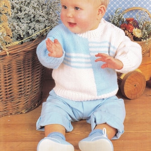 May include: A baby wearing a blue and white striped knitted sweater with a blue and white striped hat. The baby is sitting on a wooden floor.