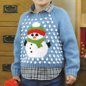 May include: A light blue knitted sweater with a snowman design. The snowman has a red hat, green scarf, and black coal eyes and buttons. White bobbles are scattered across the sweater. The sweater is worn over a plaid shirt.