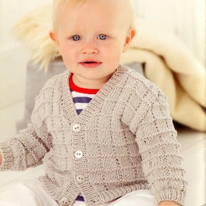 May include: A light beige knitted cardigan with a button fastening. The cardigan has a V-neck and a textured pattern. The cardigan is being worn by a young child.